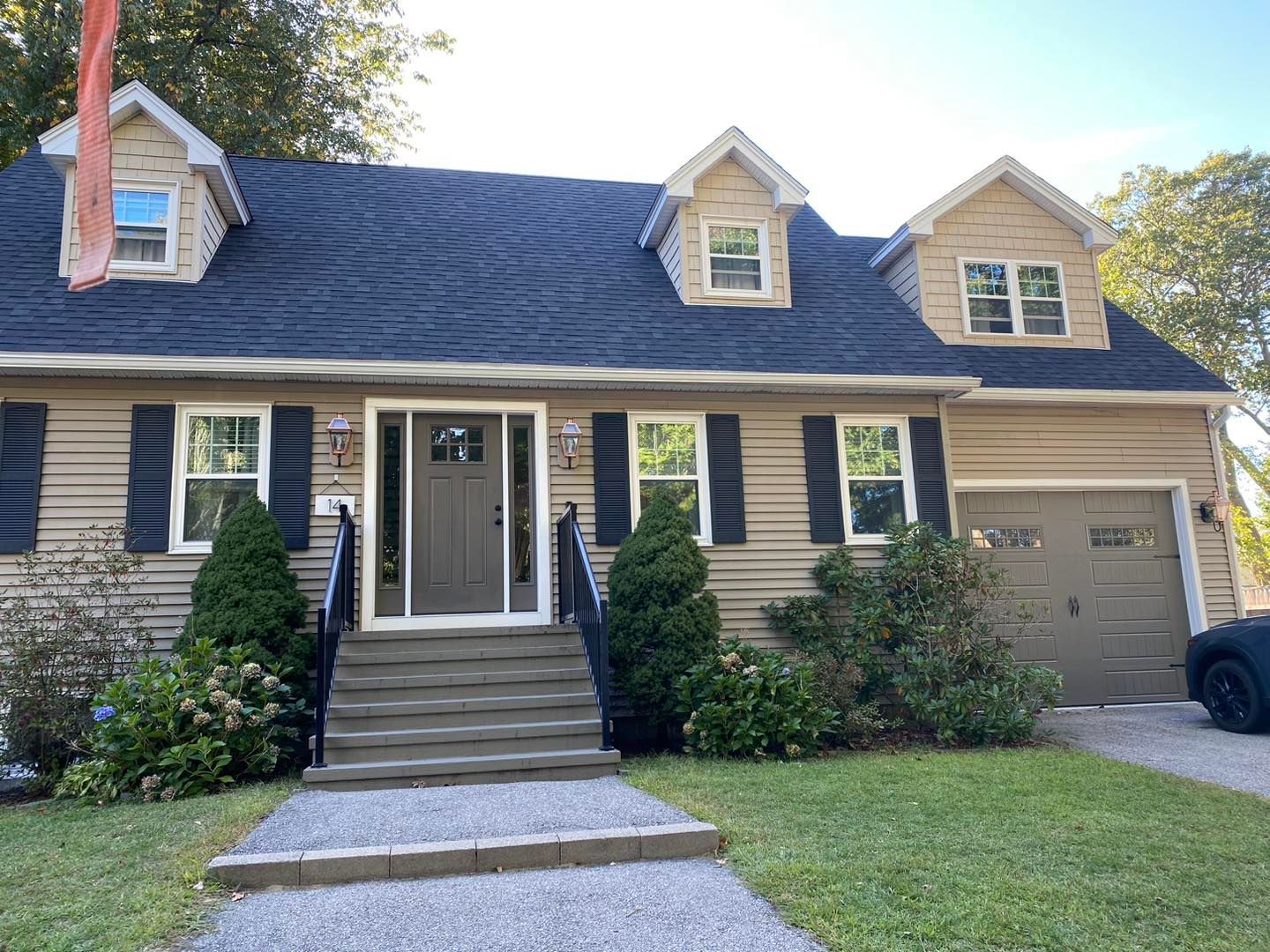 Tan house with dark roof and dormers, black shutters, front steps, and garage on a sunny day.