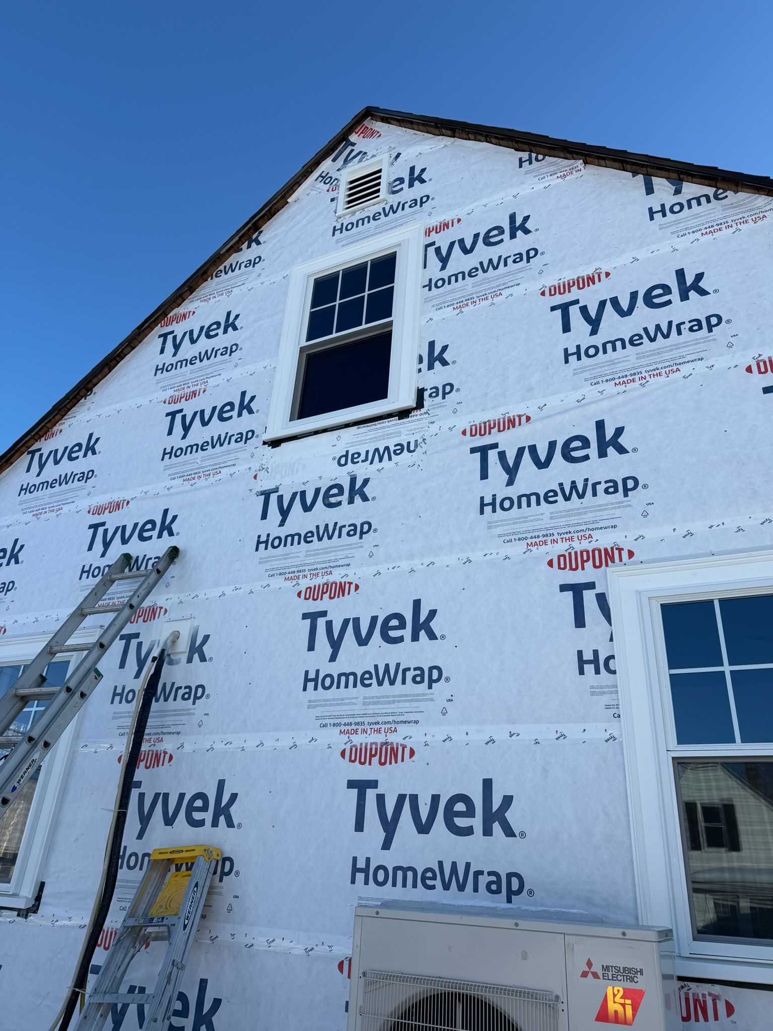 House under construction wrapped in Tyvek, with a ladder leaning against the wall under a blue sky