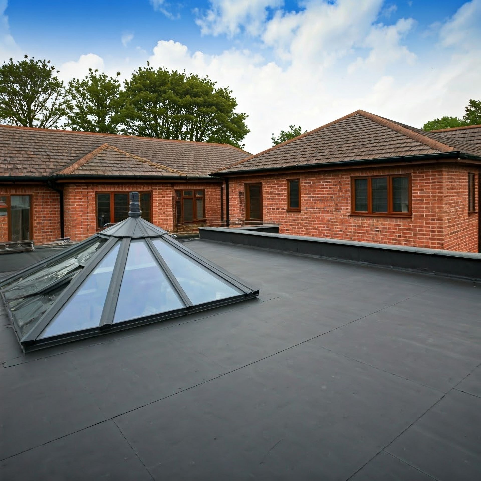 Flat black roof with a glass skylight, surrounded by red brick buildings and trees under a partly cloudy sky