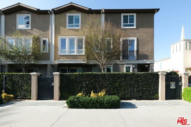 Multi-story townhouses behind a green hedge and gated entrance. Blue sky.