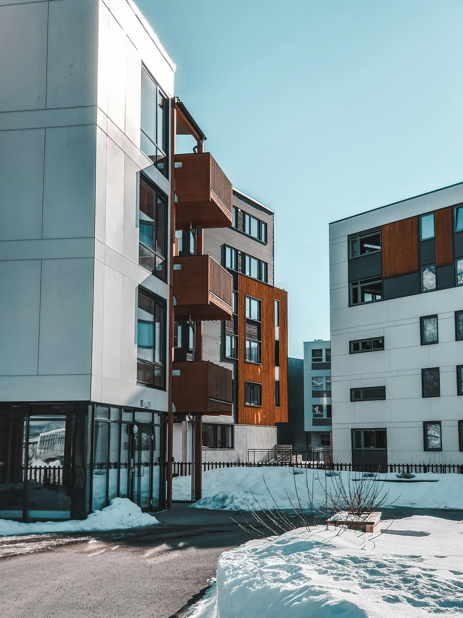 Modern apartment buildings, white and brown, with snow on the ground, against a clear blue sky.