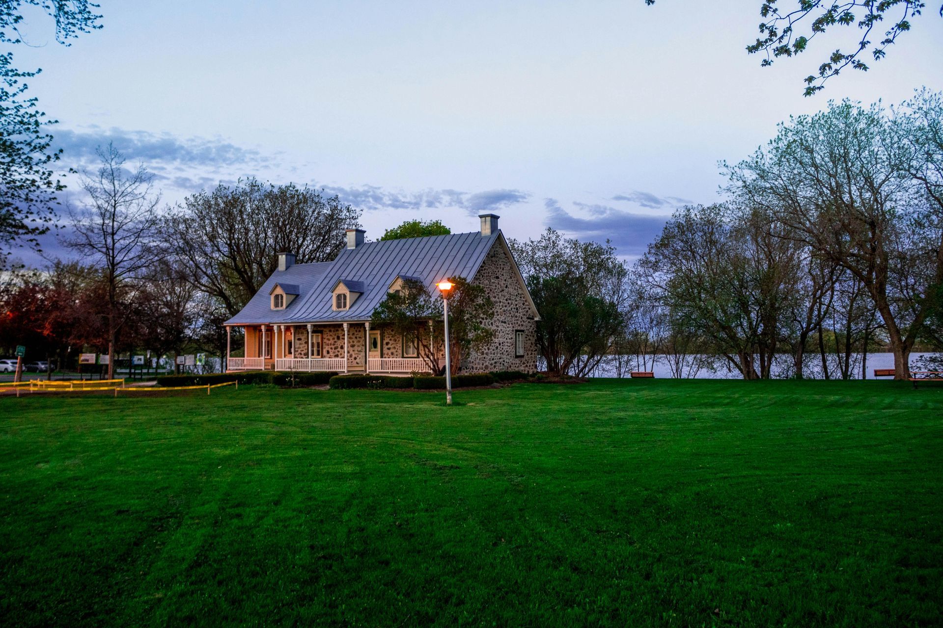 Stone house with a gray roof and dormer windows on a green lawn near a body of water, dusk.