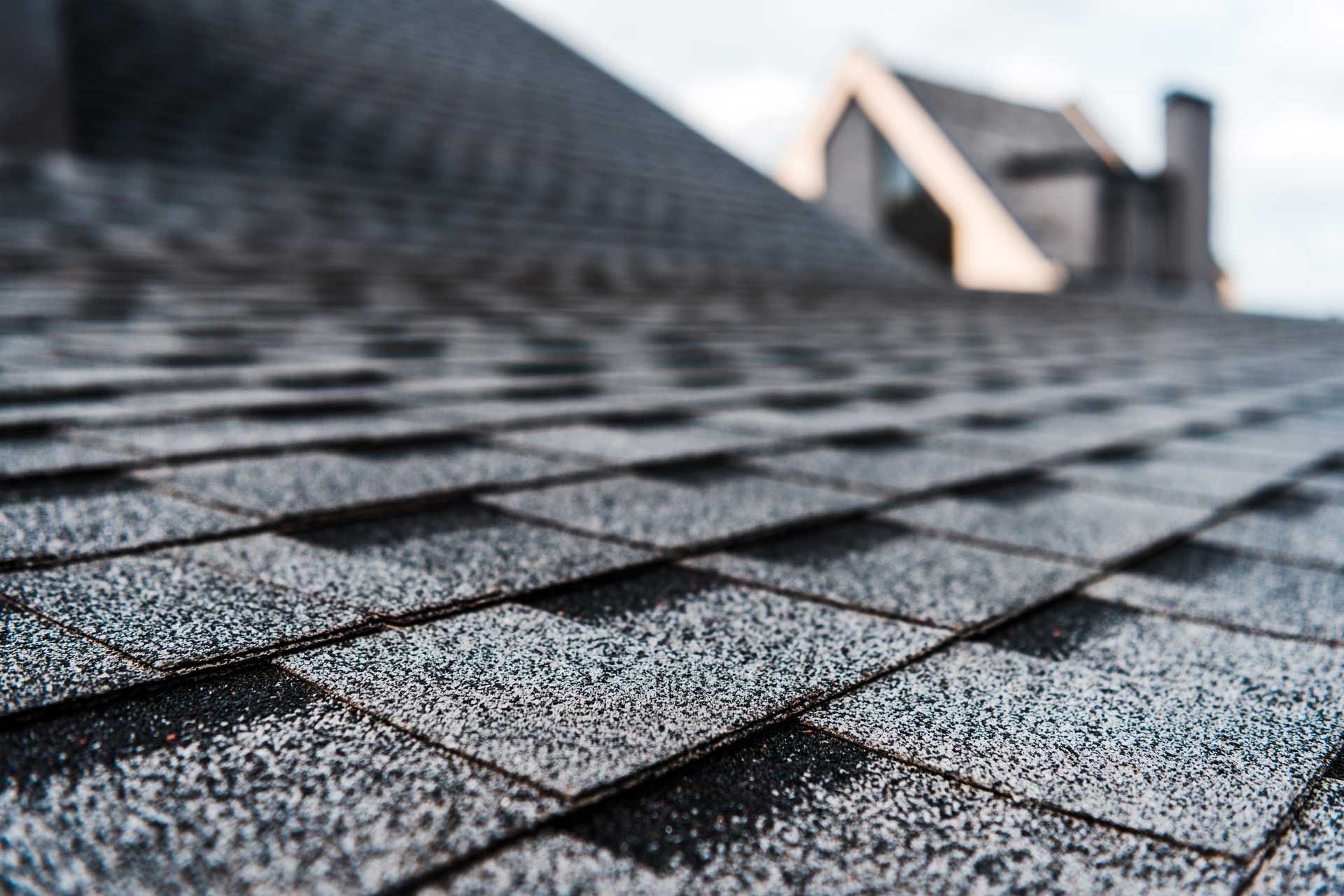 Close-up of a gray asphalt shingle roof, with a blurred house in the background.