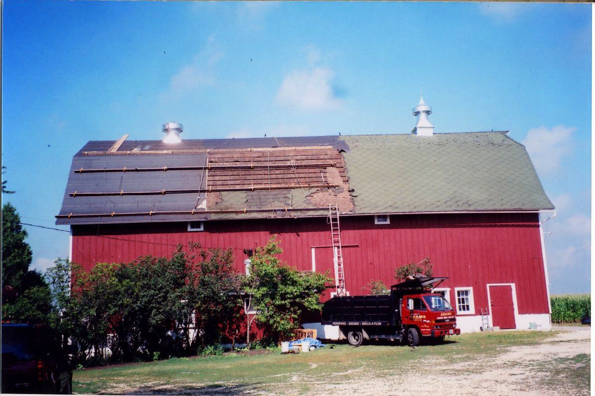Red barn with a partially replaced roof. A red truck is parked nearby. Blue sky.