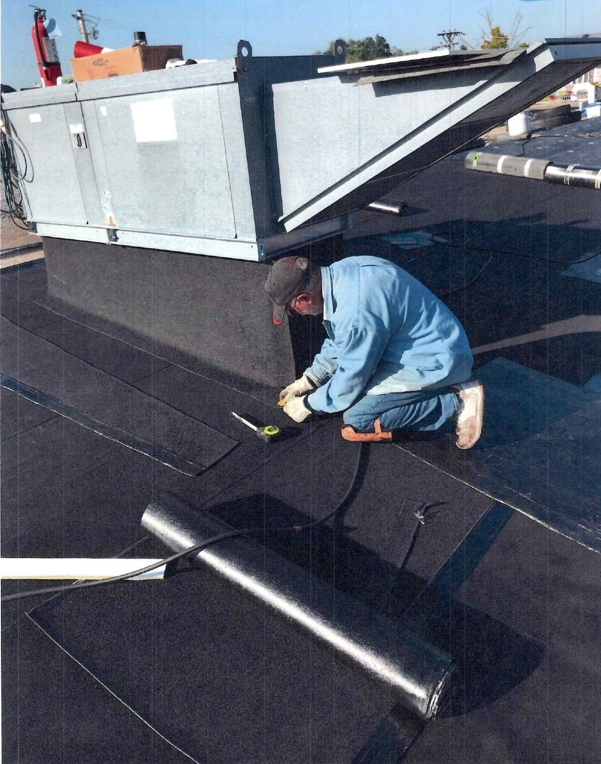 Person in blue jumpsuit working on a flat, black commercial roof near HVAC unit.