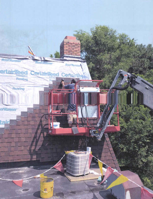 Two-story beige house with a gray roof and attached garage; flowers and green grass in front under a blue sky.