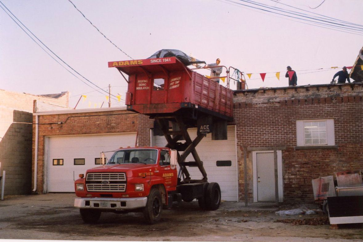 Red dump truck with raised bed next to a brick building; people on the roof.
