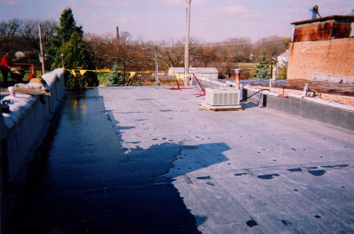 Flat rooftop with a dark, wet coating; a brick building is visible.