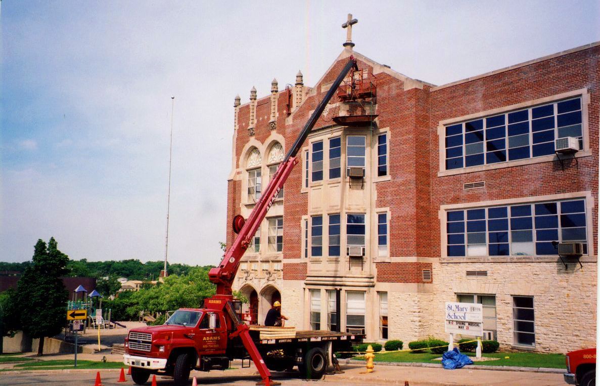 Red crane platform next to a brick building; cross on top. Workers are visible, blue sky.
