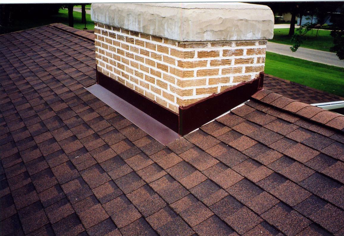 Brick chimney on a brown shingled roof with dark metal flashing.