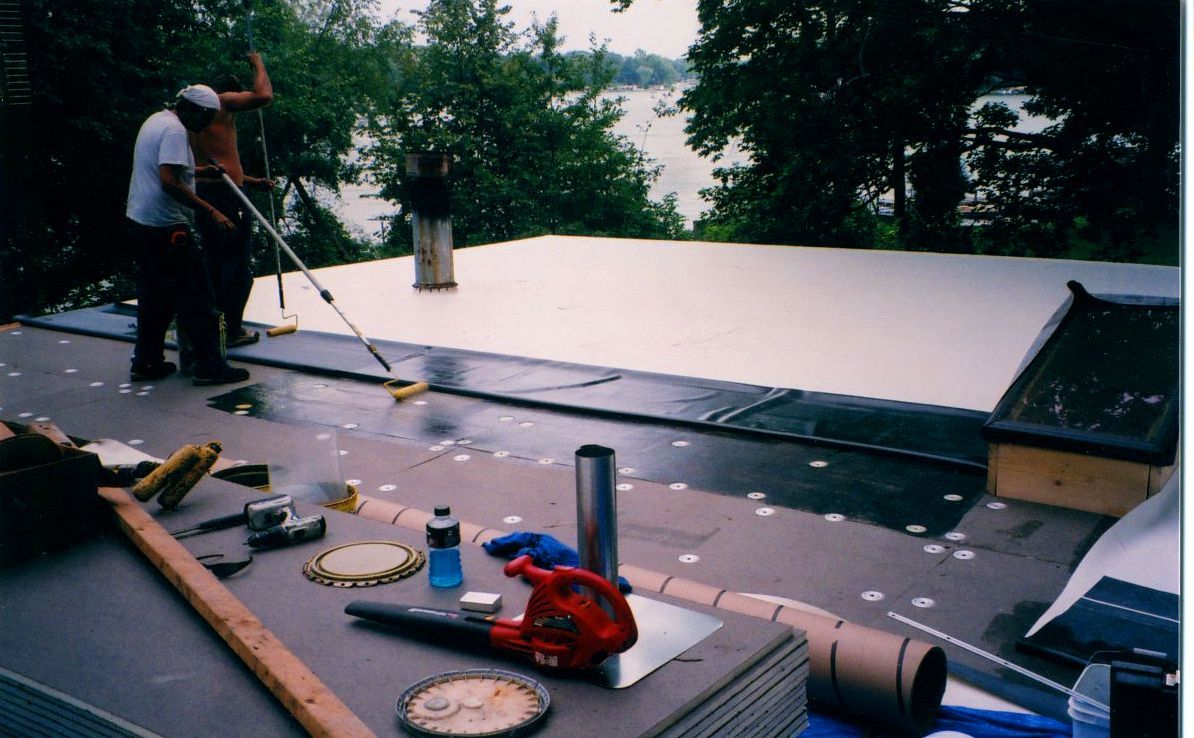Two people install a white flat roof. One uses a roller. Tools and materials are scattered on the roof.