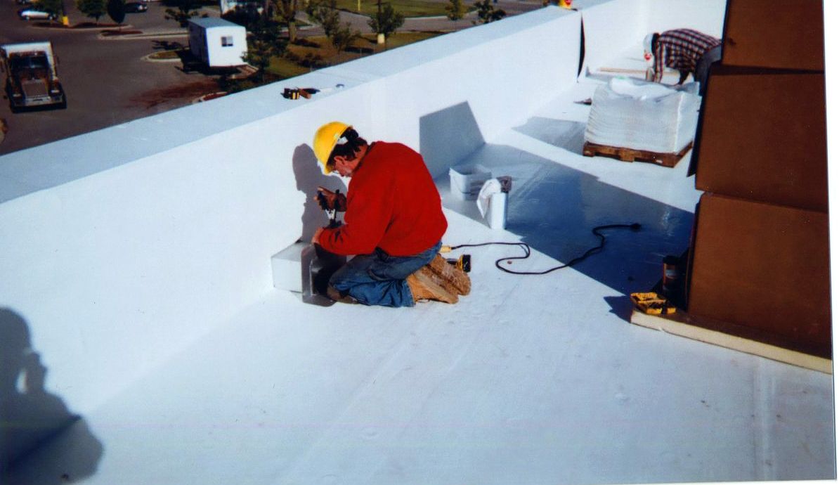 Person in yellow hardhat and red shirt working on a white flat roof. Building materials and a truck are visible.