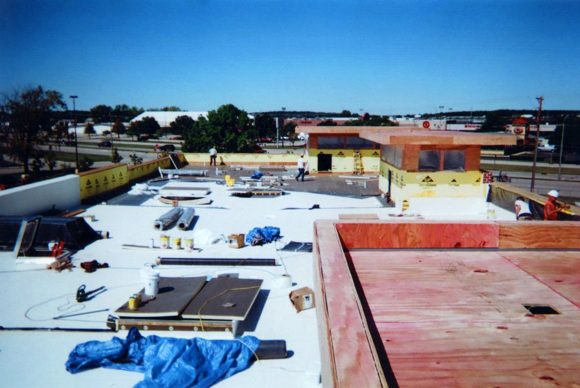 Construction workers on a flat roof. Tools and materials are scattered, a building is under construction, and the sky is blue.