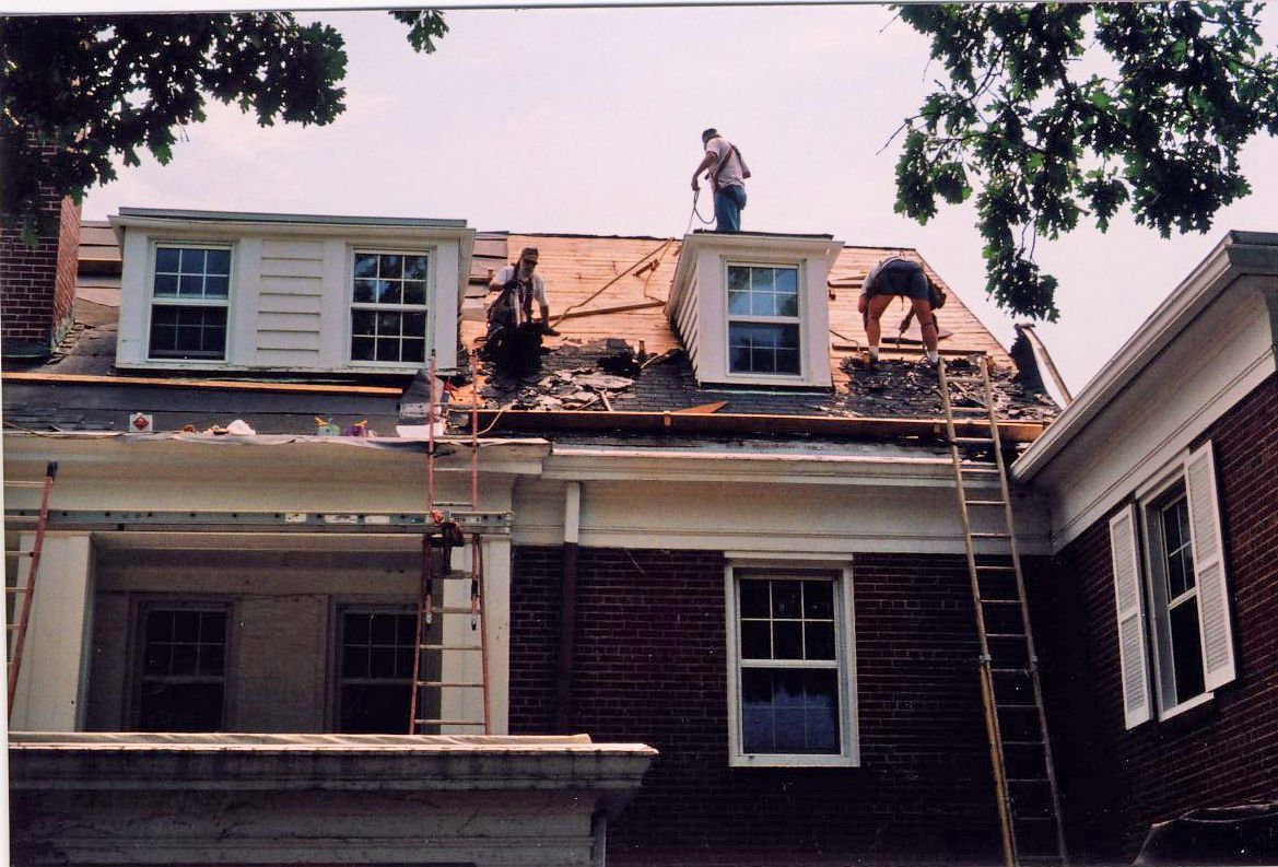 Construction workers on a roof, removing old shingles. Brick building with white trim, ladders, and trees.