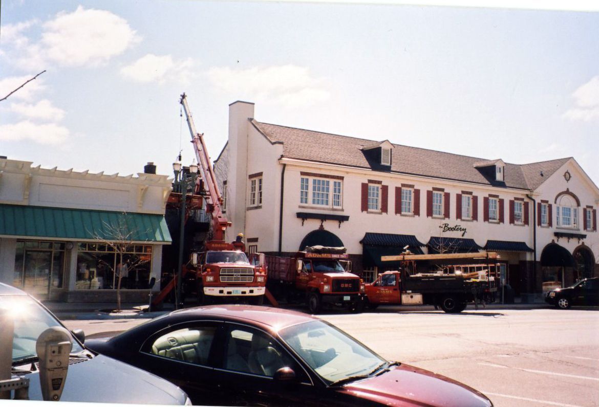 Trucks with a crane working on a building. White building with black awnings, red shutters, blue sky.