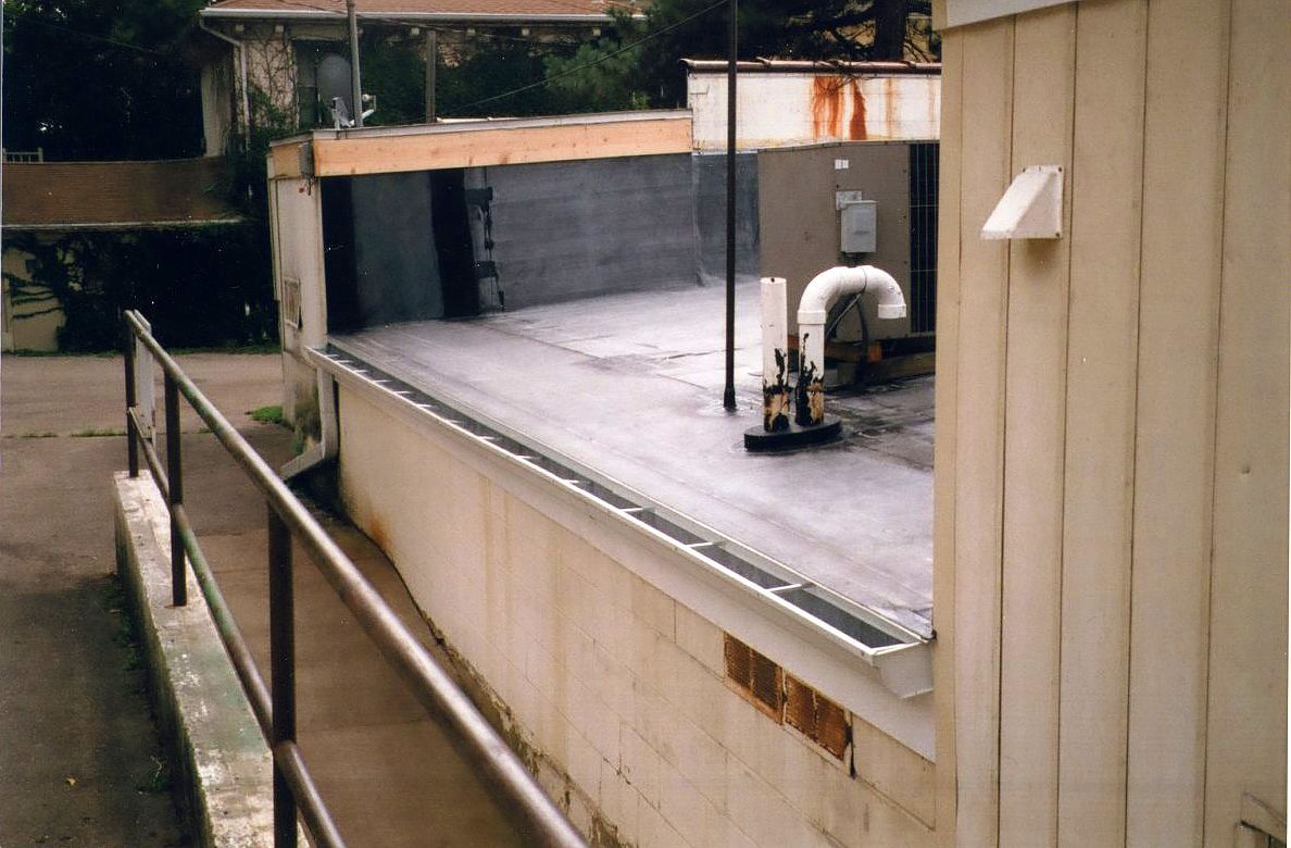 Rooftop with a railing, featuring a flat black surface, pipes, and a building's white siding.