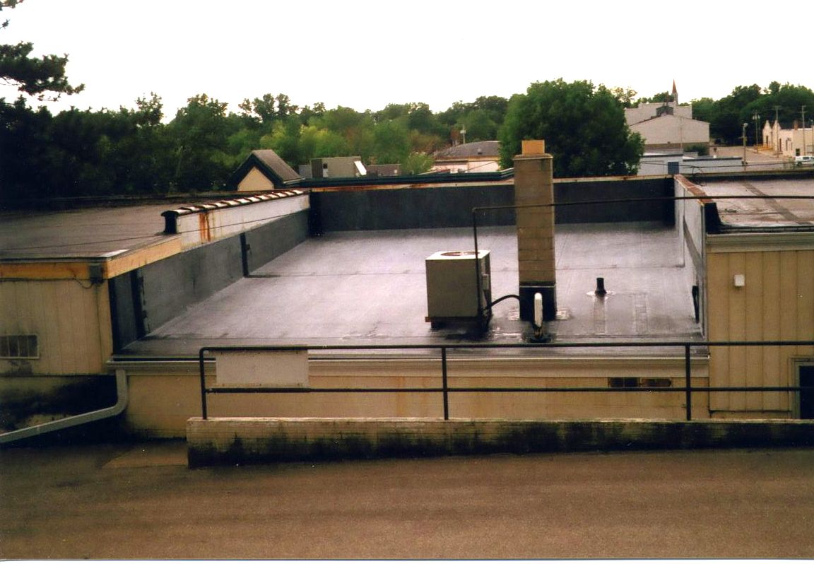 Flat roof with HVAC unit and chimney, bordered by a low wall and railing. Trees and buildings in the background.