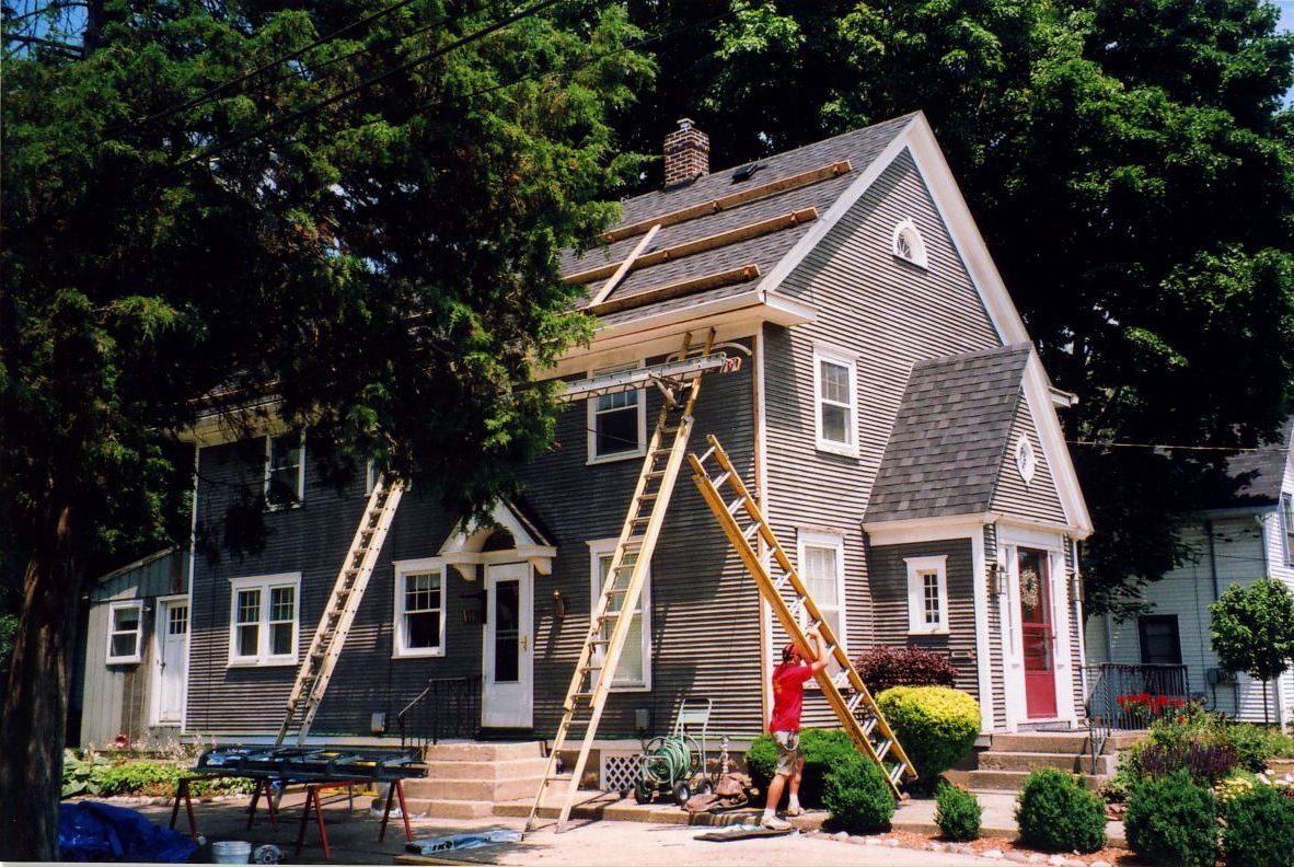 House with ladders and roof work in progress. Person on one ladder, gray siding. Trees and bushes in yard.