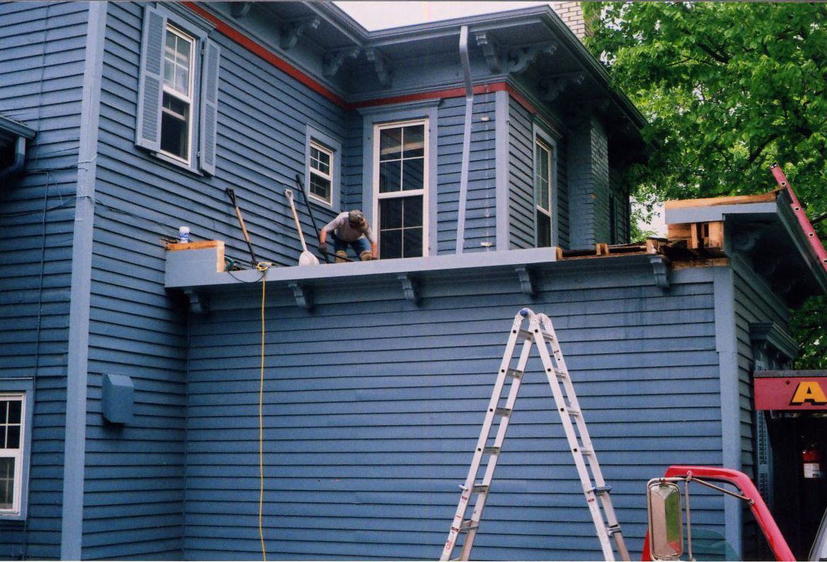 Person working on exterior of blue house, standing on a low-roofed section with ladder and tools.