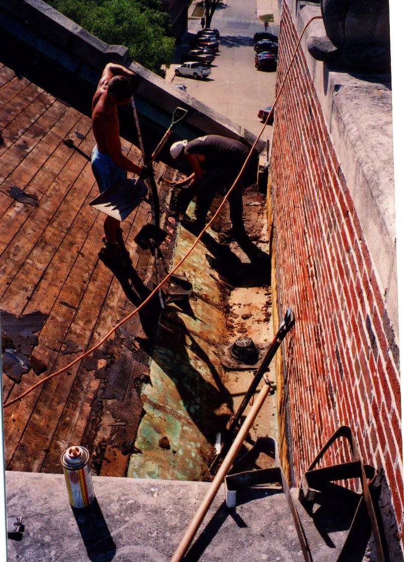 Two people working on a roof next to a brick wall; rusty metal and wooden planks are visible.