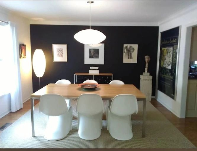 Modern dining room with white chairs around a light wood table, black accent wall, art, and a large pendant light.