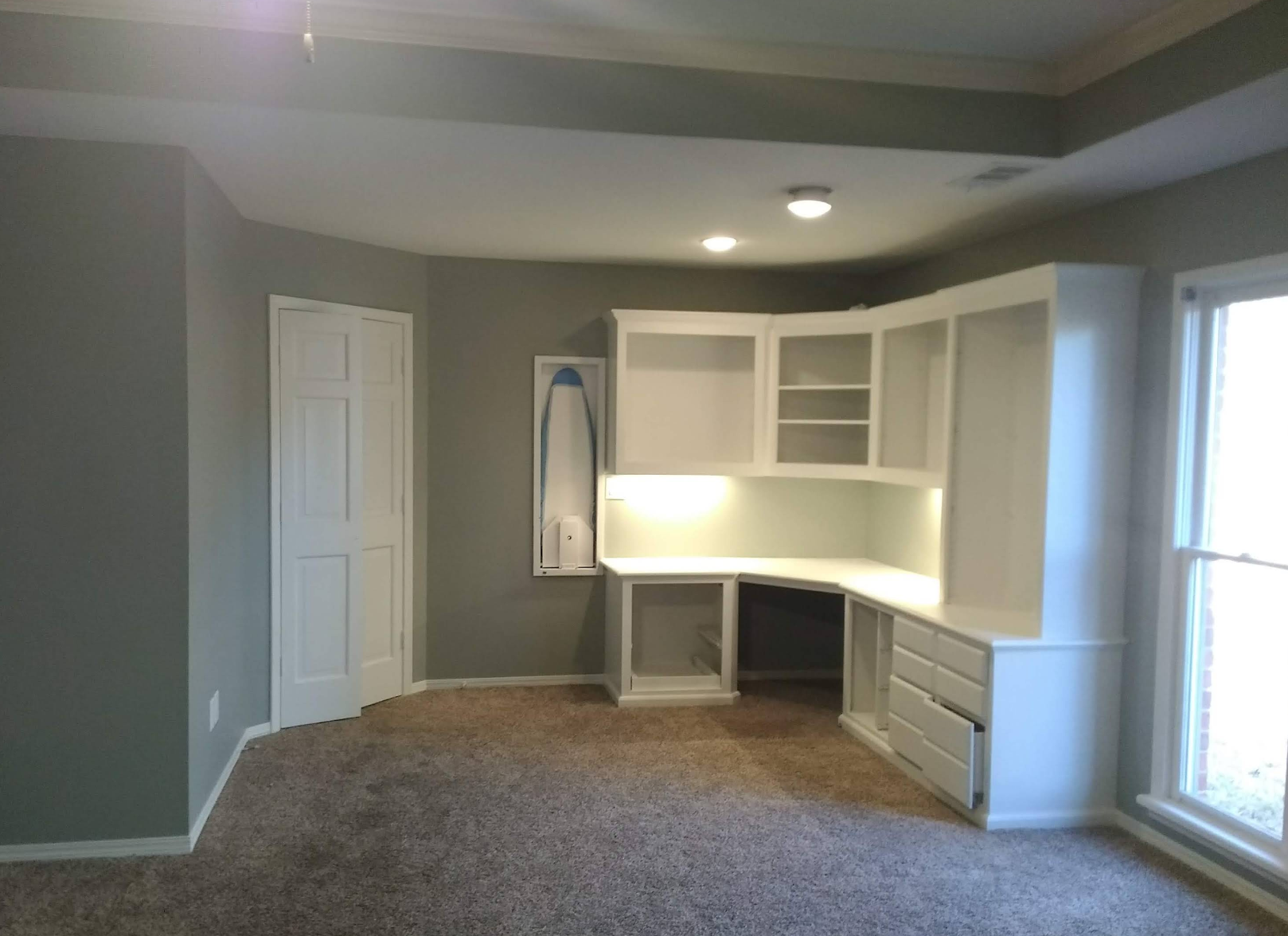 A room with a white built-in desk and cabinets, a door, and a window. Light brown carpet, gray walls.
