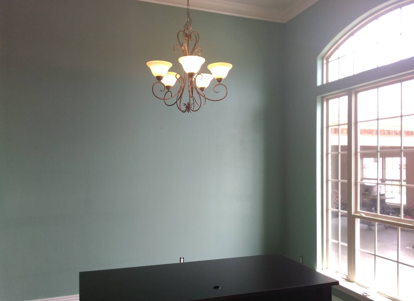 A room with blue-green walls, chandelier, a black desk, and a large window.