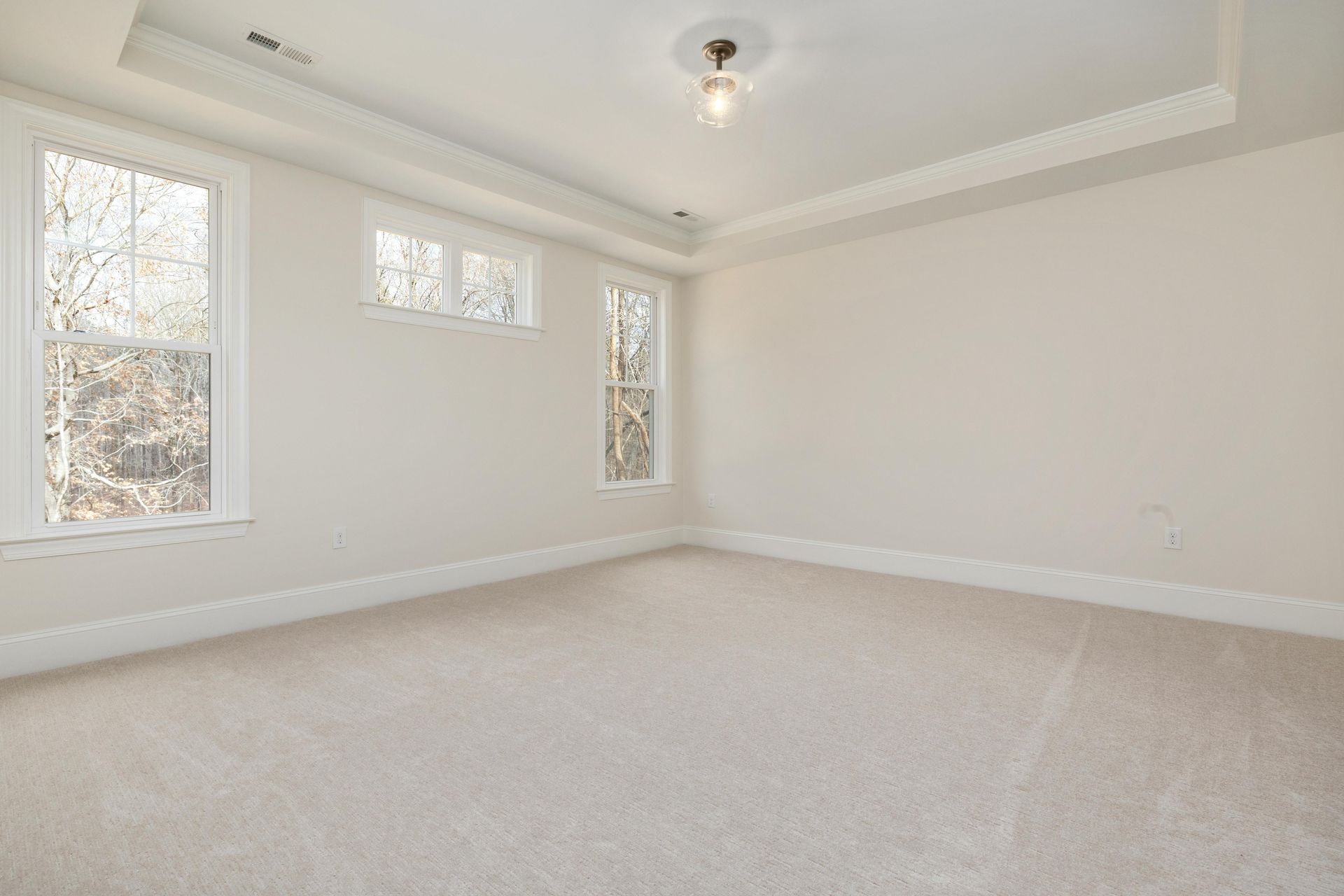 Empty room with beige carpet, white walls, and three windows. A light fixture hangs from the ceiling.