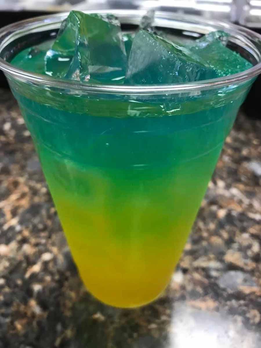 Layered, colorful drink in a clear plastic cup with ice cubes. Yellow, green, and blue gradient.