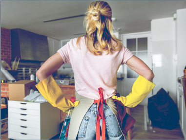 一位戴著手套和圍裙的女子站在廚房裡，雙手叉腰，看著凌亂的房間。