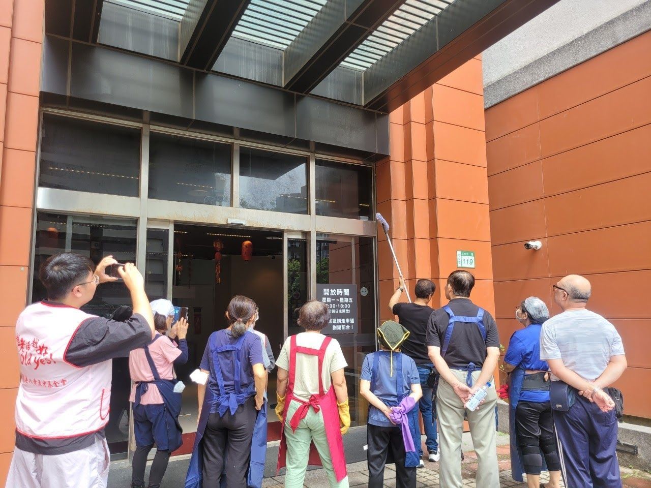 人們用長柄刷子清潔建築物入口；其他人則在一旁觀看並拍照。