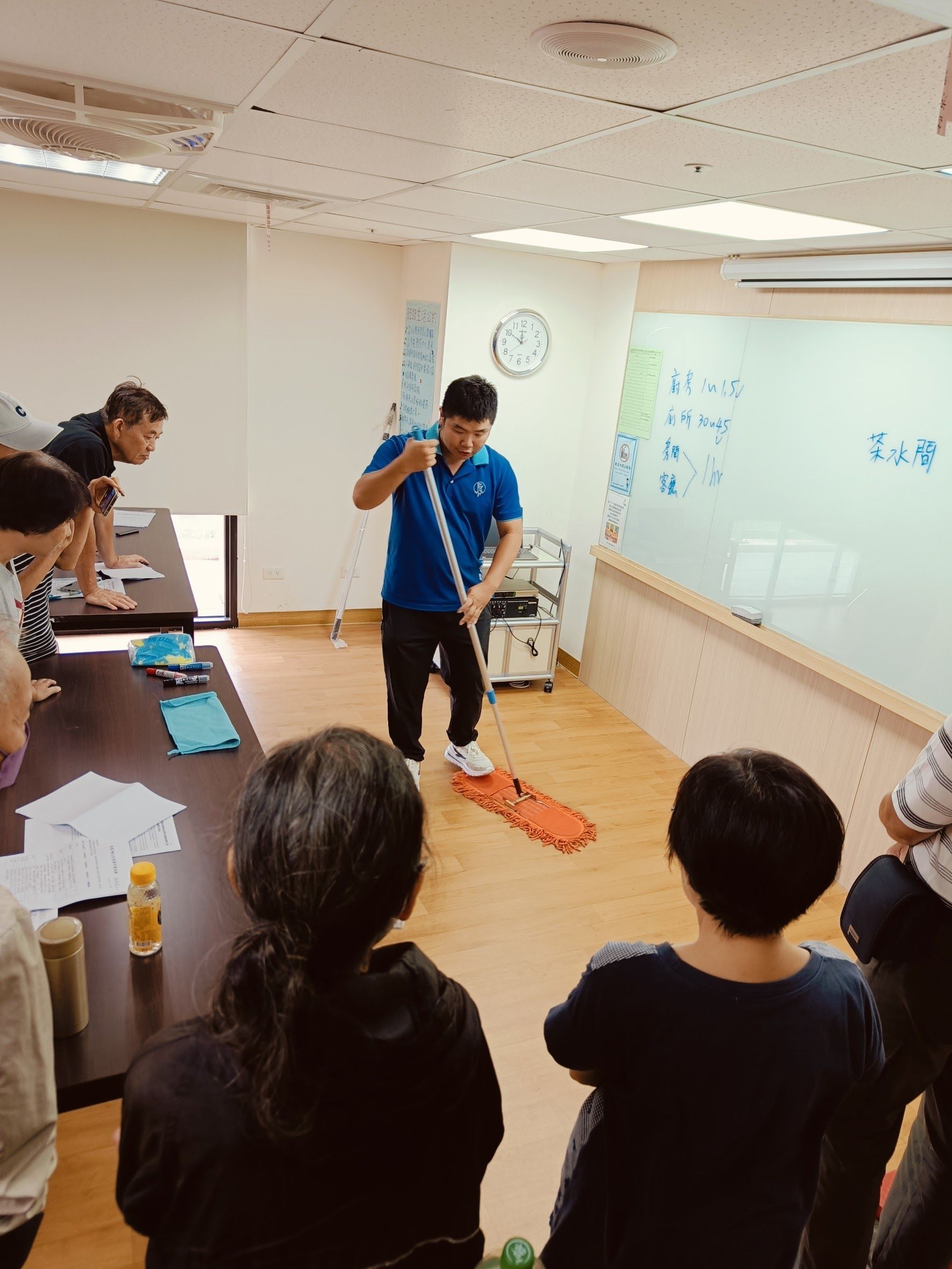 一名男子在教室裡用拖把示範。學生們觀看他在教學環境中拖地。