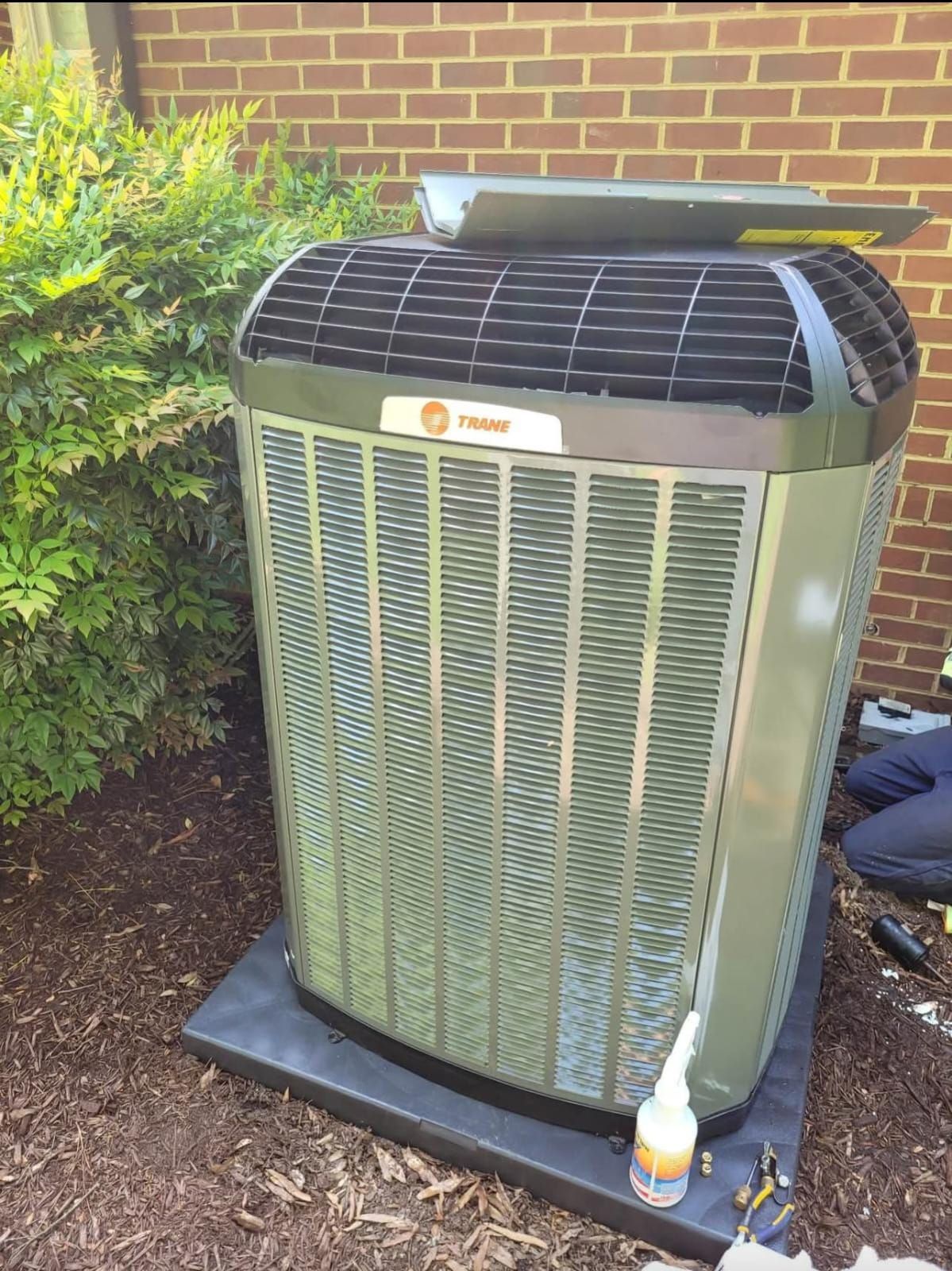 A Trane air conditioner unit near a brick wall and bushes, on a black pad.