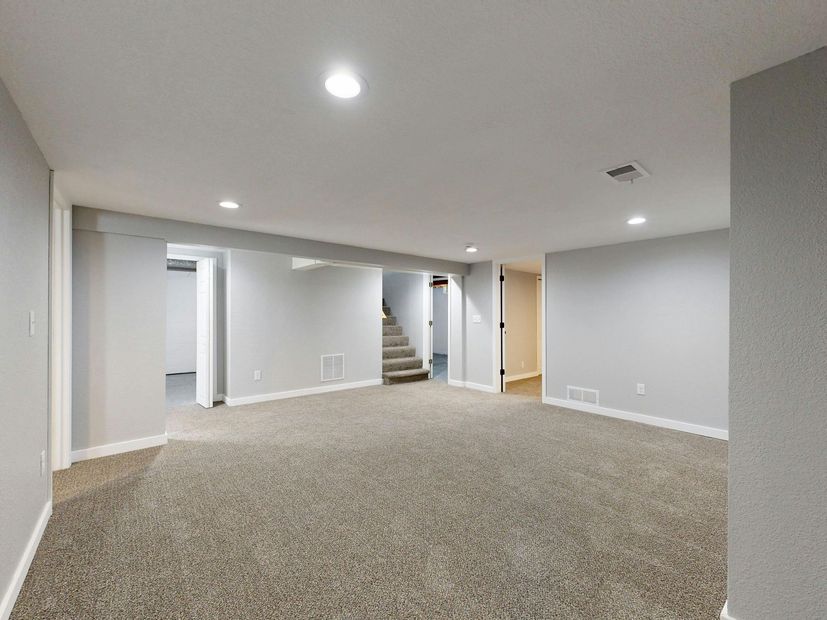 A wide shot of a finished basement with light gray walls, brown carpet, recessed ceiling lighting, and a staircase.
