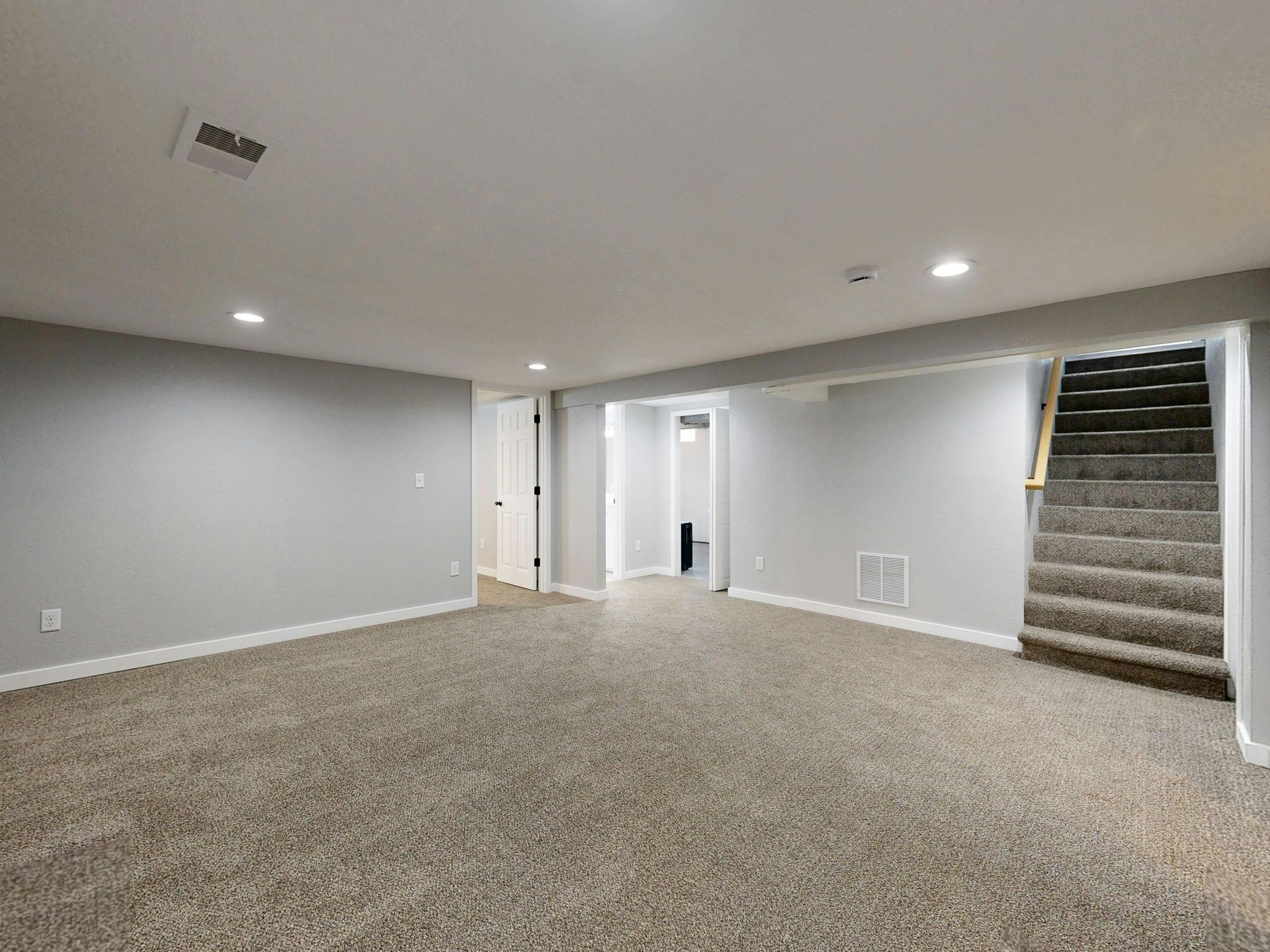 A basement room with light gray walls, neutral speckled carpeting, recessed lighting, and stairs leading upward.