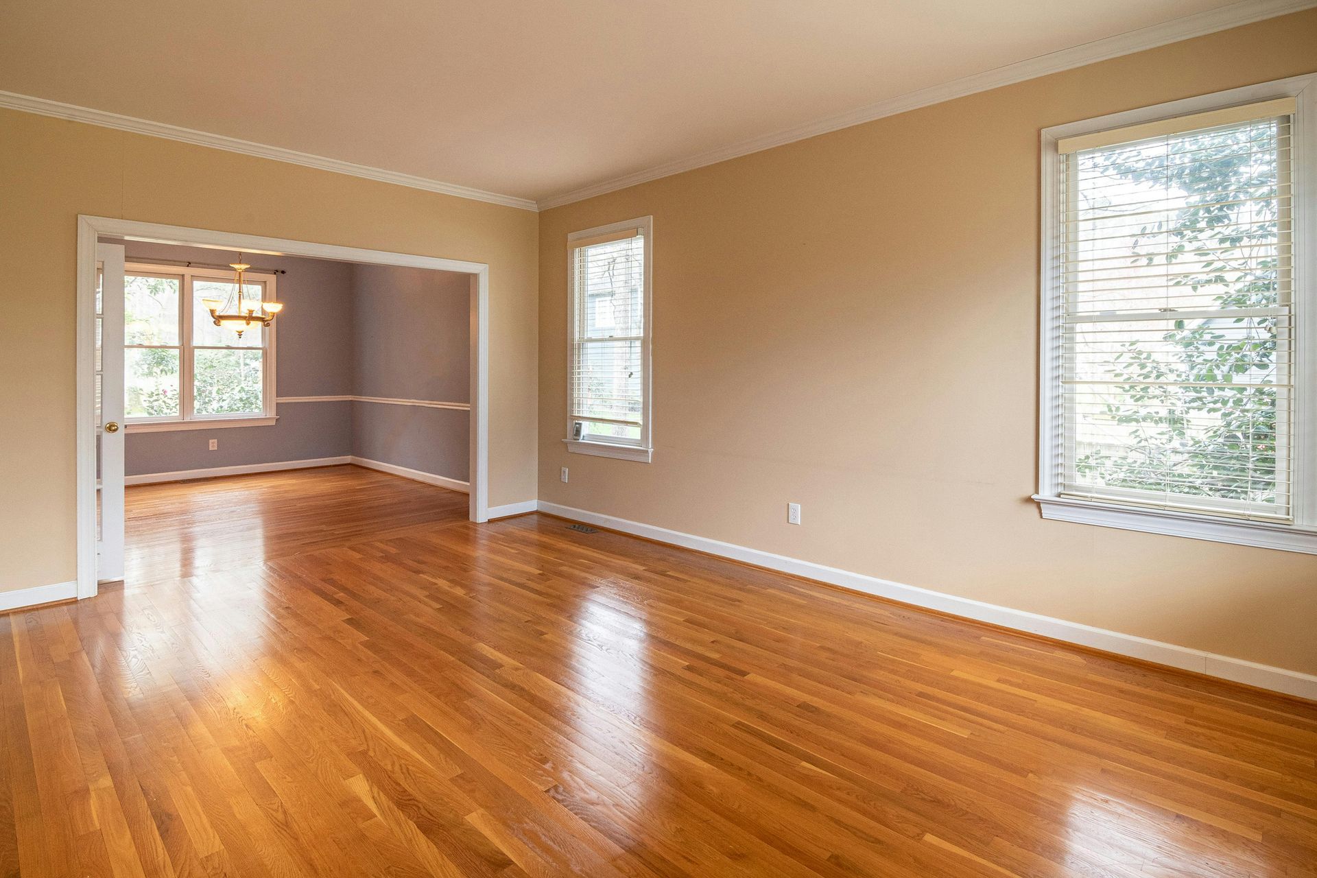 A spacious room with polished hardwood floors, beige walls, and a large window, leading to a connected dining area.