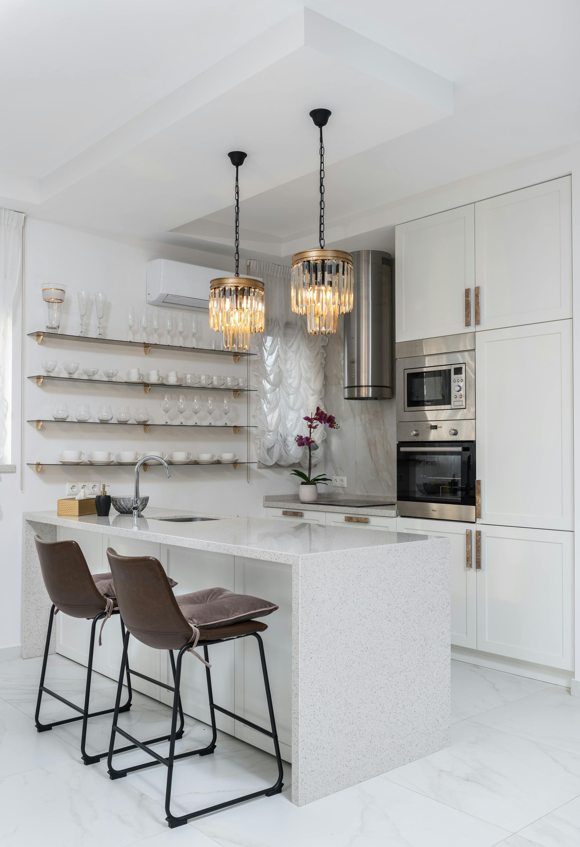 A modern white kitchen with a speckled stone island, two brown bar stools, glass pendant lights, and open shelving.