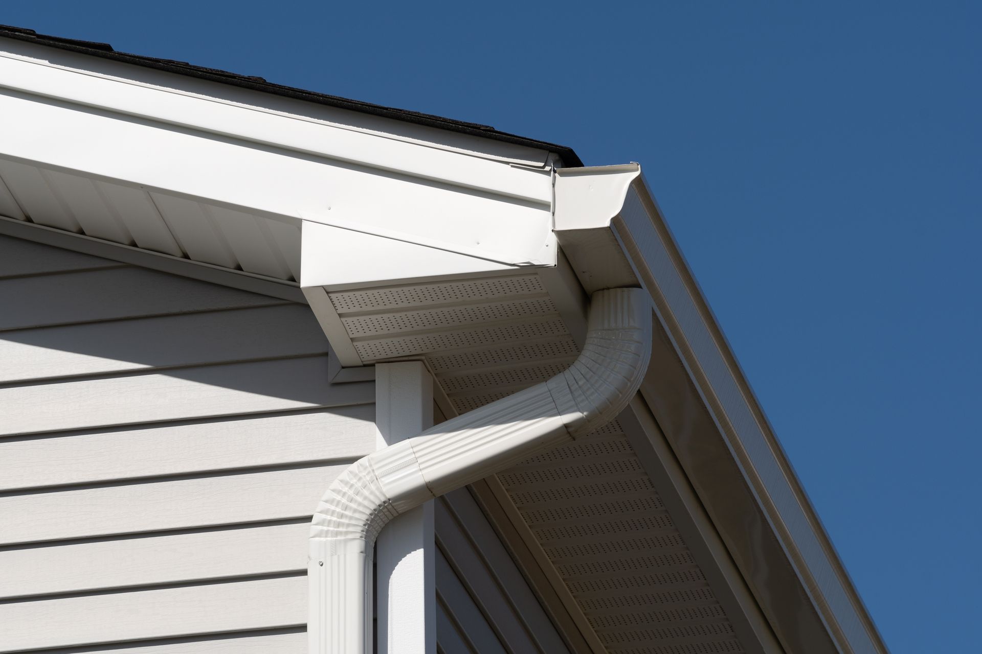 White house siding and roof eaves with a white rain gutter downspout against a clear blue sky.