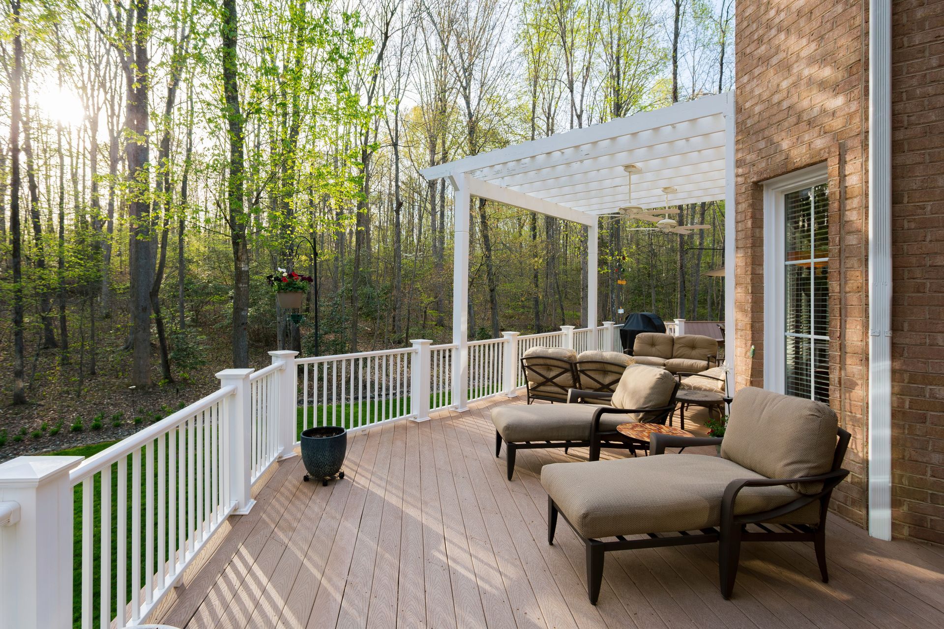 A sunny backyard deck featuring tan lounge chairs under a white pergola, with white railings overlooking a wooded area.