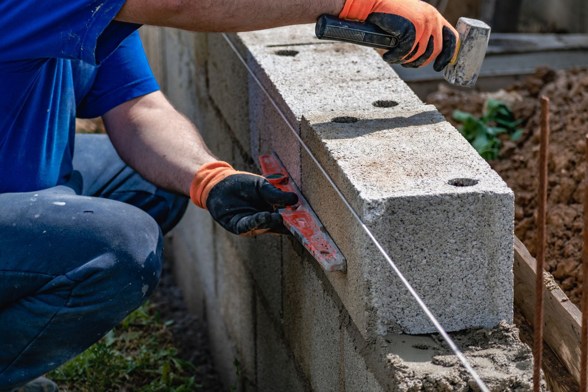 A person in a blue shirt and work gloves uses a mallet and a level to lay a concrete block on a wall.