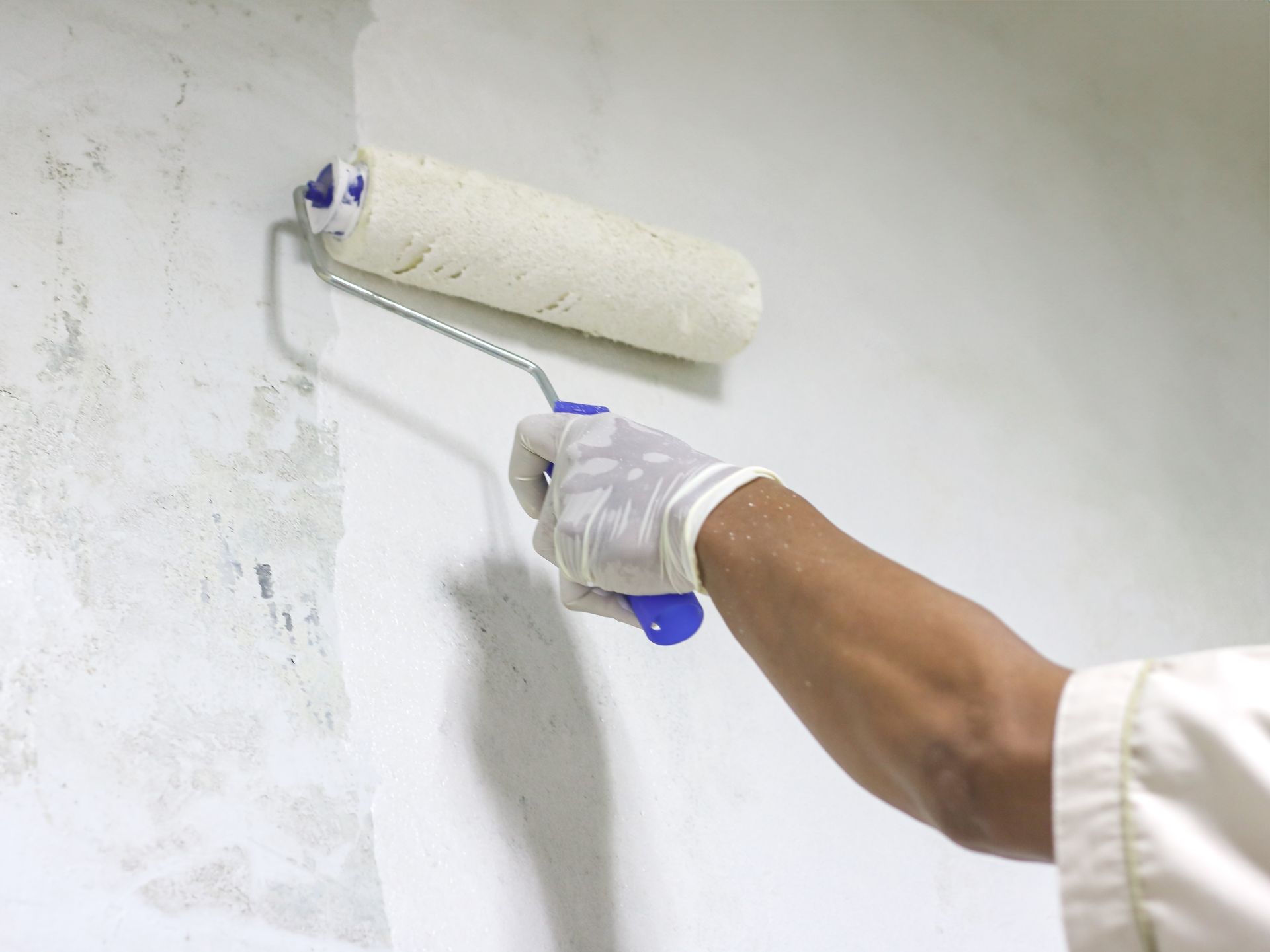 A person wearing a white glove paints a wall white with a roller.