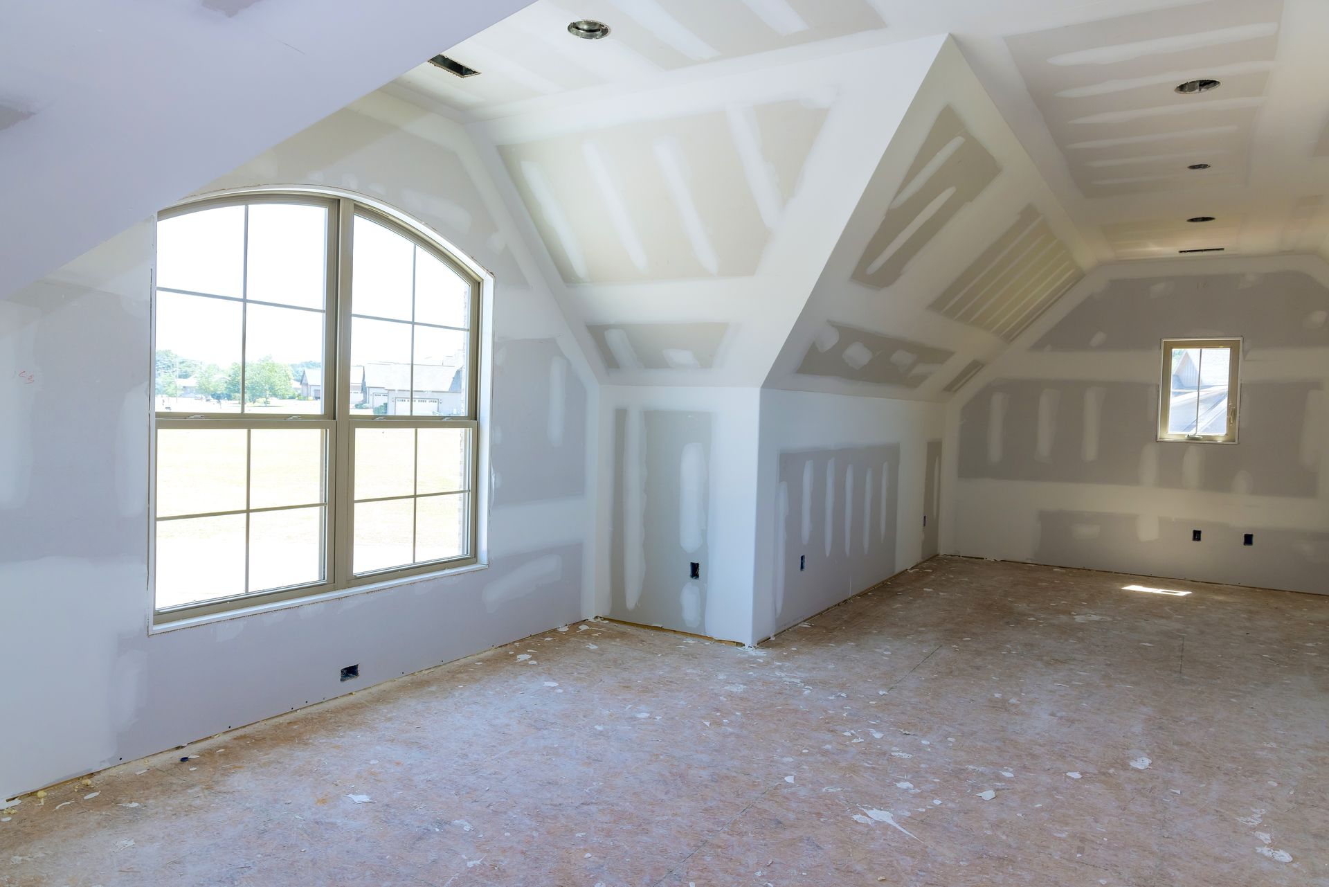 An unfinished attic room featuring newly installed drywall, an arched window, a smaller window, and an unfloored subfloor.