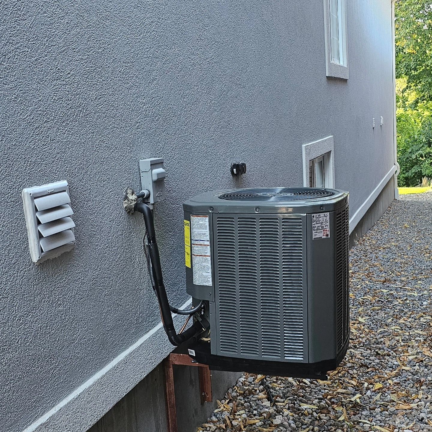 Air conditioner unit outside gray building with vent and gravel.