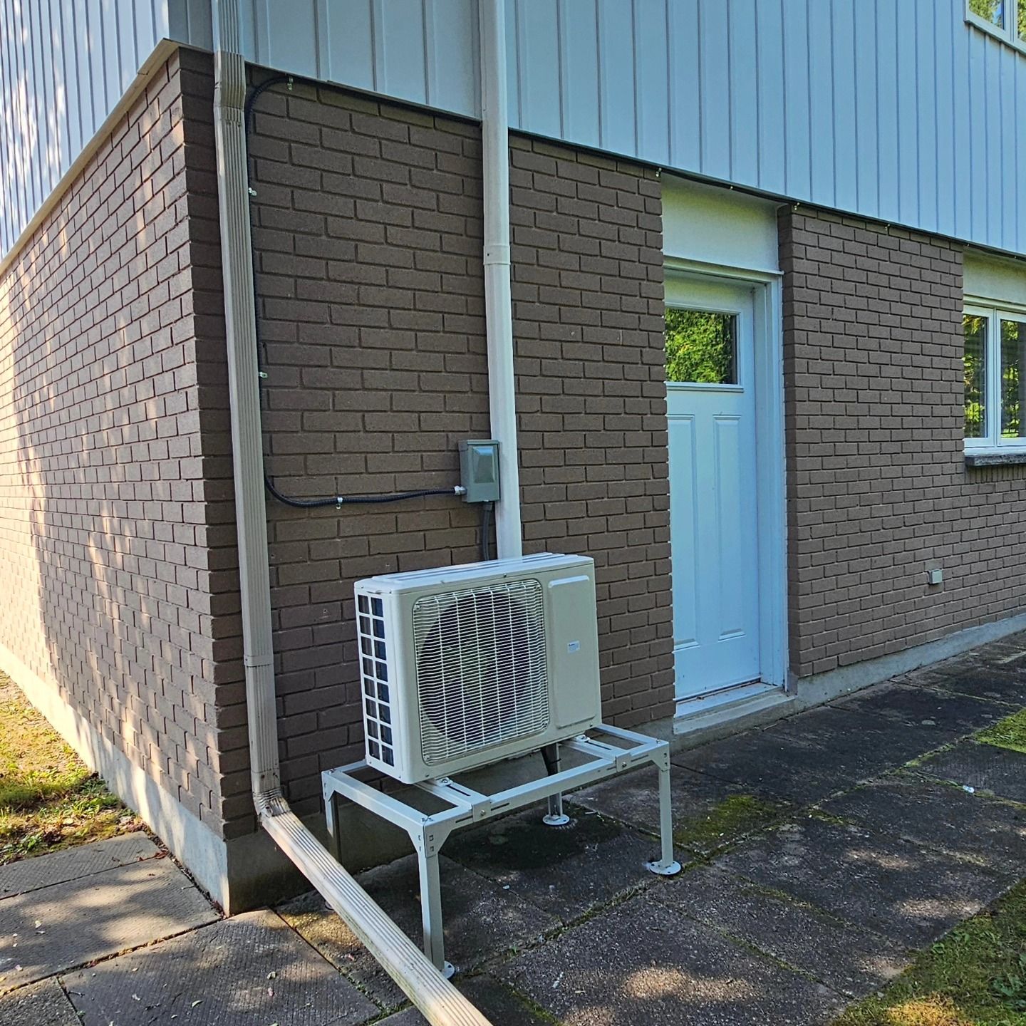 Air conditioning unit on a metal stand outside a building with brick wall and a white door.