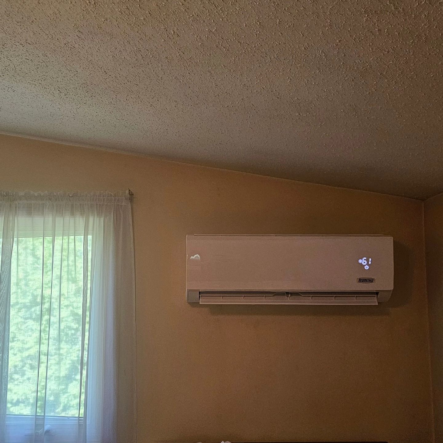 Wall-mounted air conditioner unit on a beige wall next to a window with white curtains.