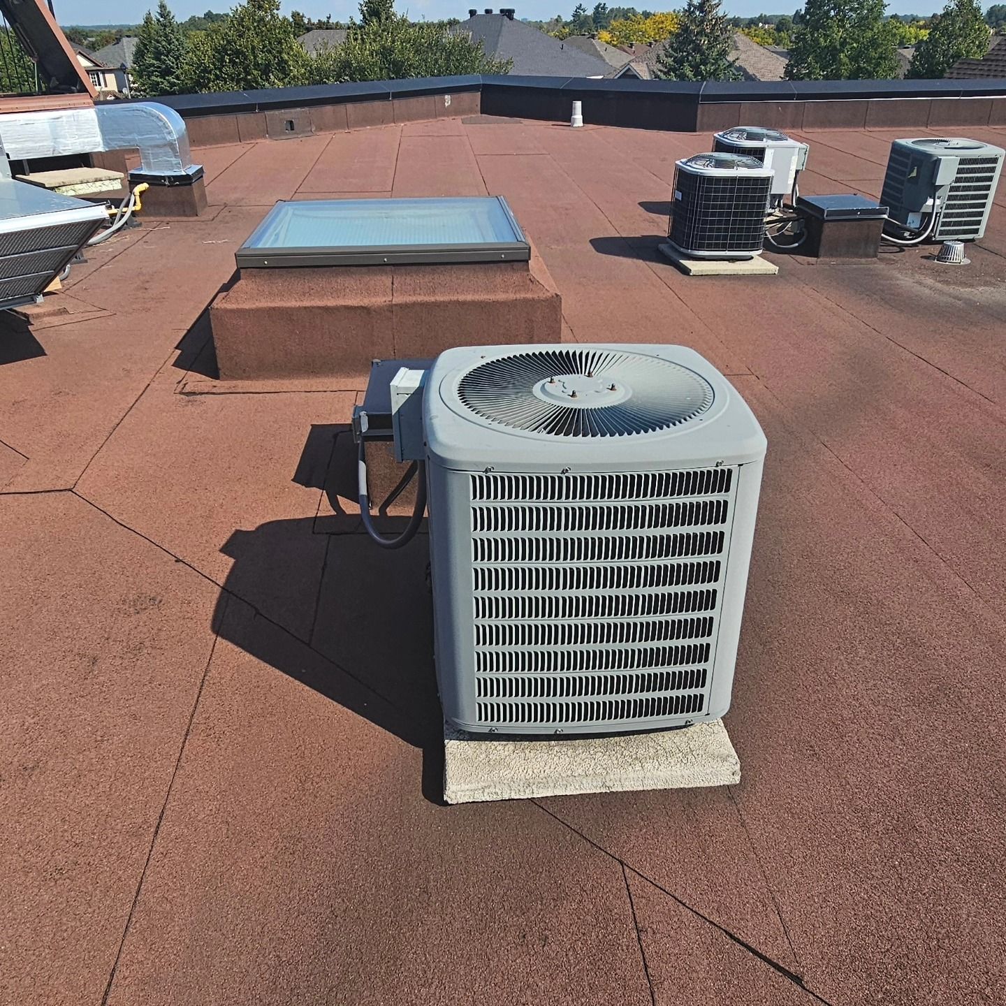 Air conditioning unit on a flat roof with other equipment and a skylight, in bright sunlight.