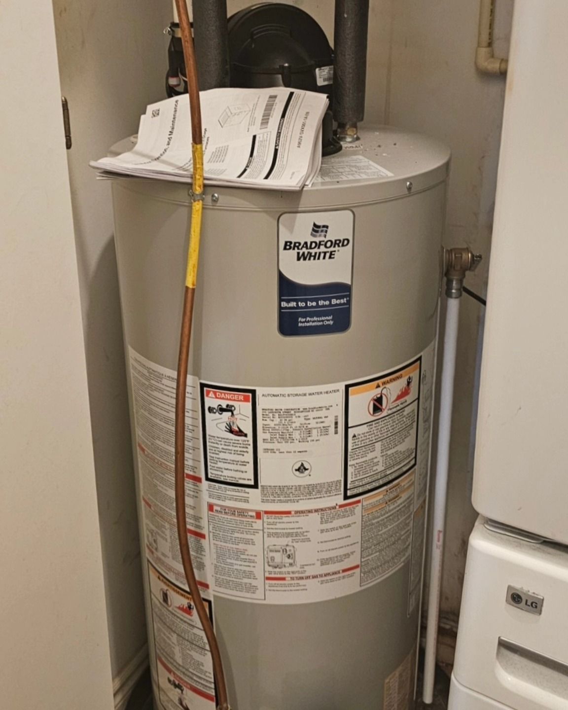 A water heater in a utility closet with a gas pipe and attached papers.