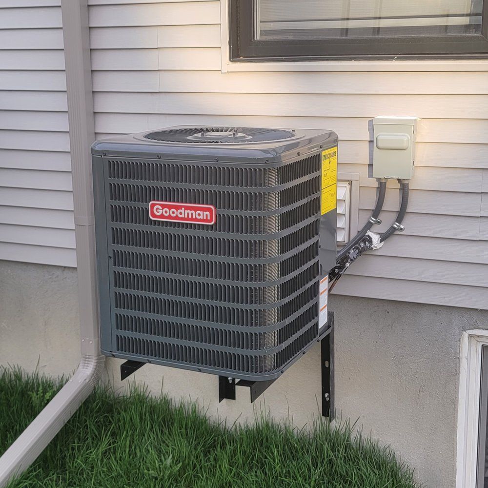 Goodman air conditioner unit mounted outside on a beige house wall next to electrical box.