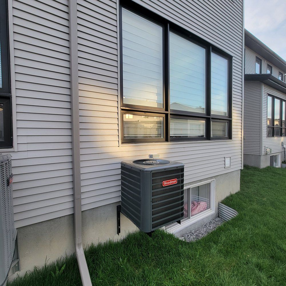 HVAC unit mounted on a grey-sided house near a window and grass. Black-framed window reflects sky.