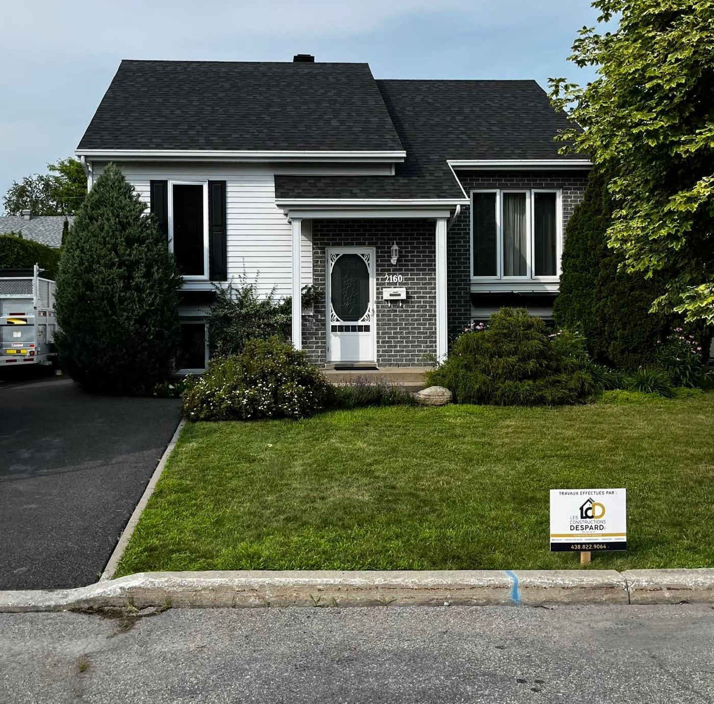 Maison blanche avec toit noir, pelouse verte, allée, buissons et panneau.